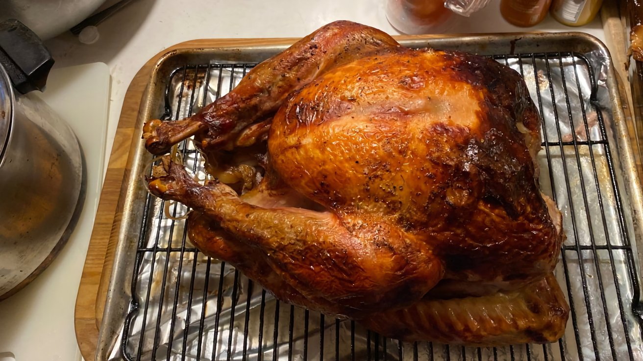 Roasted turkey with golden-brown skin on a cooling rack over a baking sheet, surrounded by kitchen items.