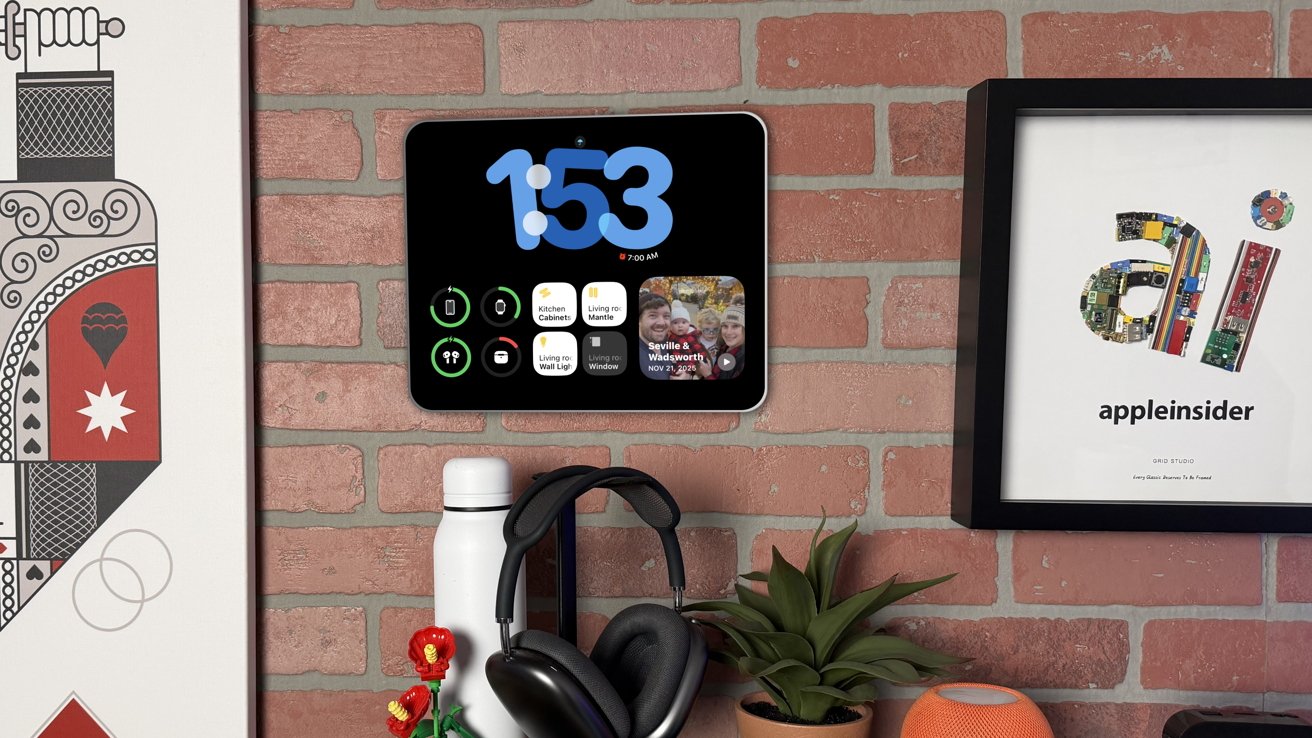 A smart display showing the time, surrounded by art, framed poster, headphones, a water bottle, plant, and a small speaker on a brick wall backdrop.