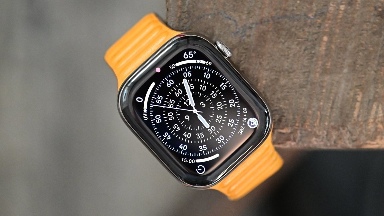 Smartwatch with orange band on wooden surface, displaying an analog watch face with white numbers and markers on circular black screen, showing temperature and UV index.
