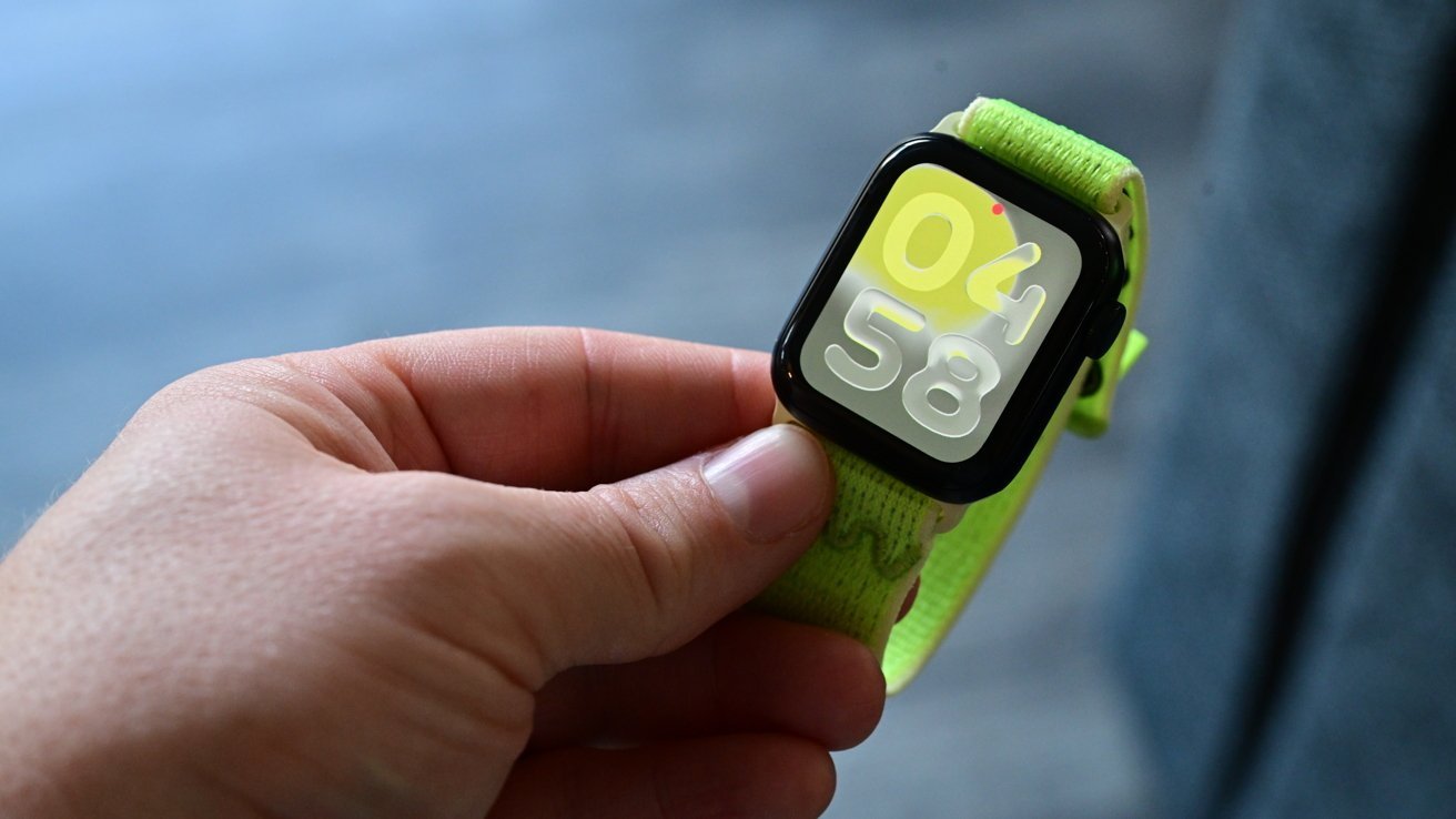 Hand holding a smartwatch with a bright green band and a digital display showing 04:58 against a blurred background.