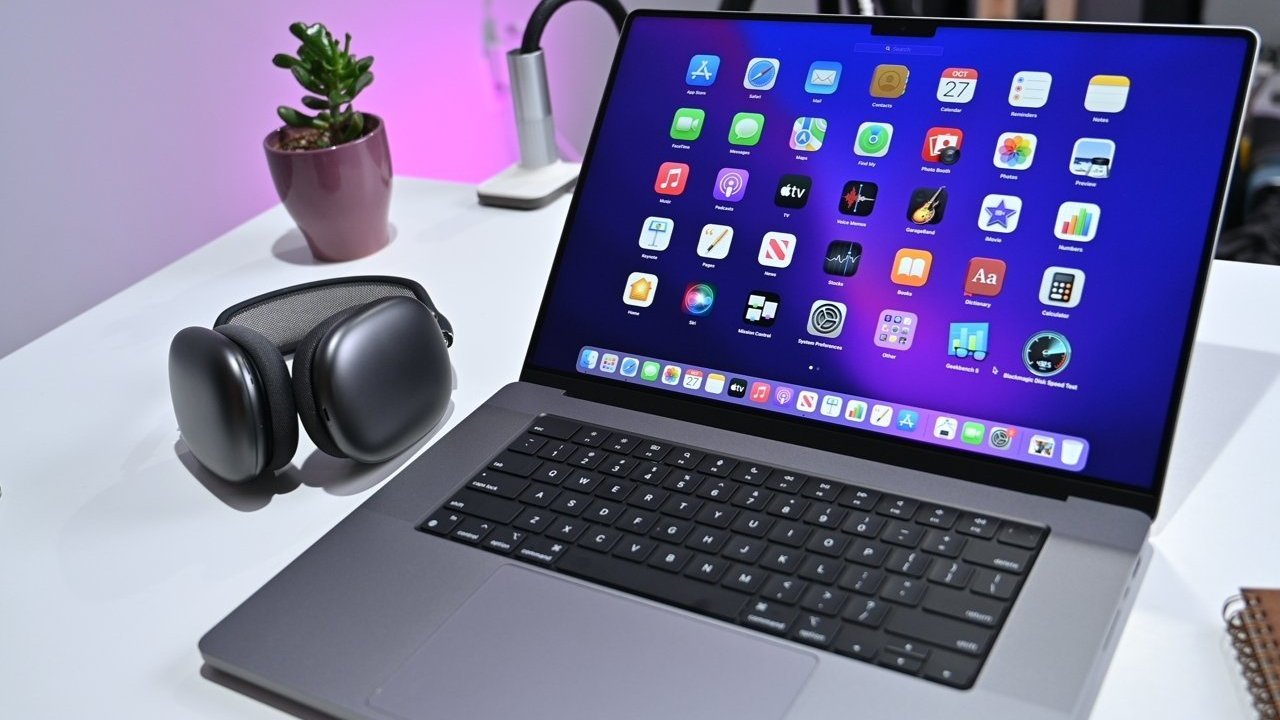Laptop displaying colorful app icons, wireless headphones, and a small potted plant on a white desk with ambient pink lighting.