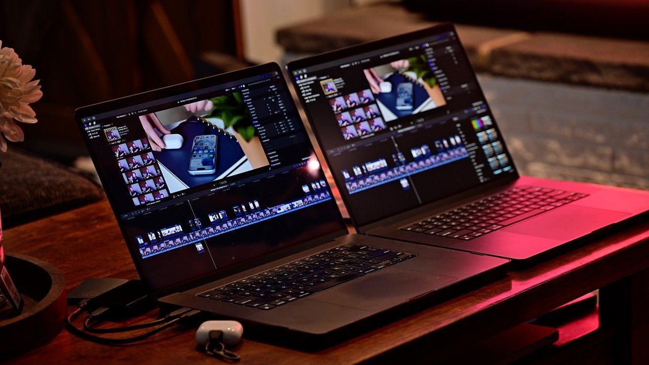 Two MacBook Pro laptops side by side on a wooden table displaying video editing software; a small white earbud case is on the table.