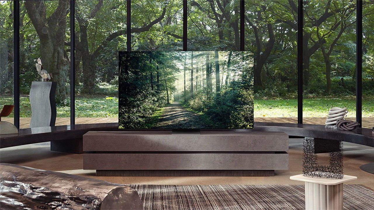 Modern living room with a large TV displaying a forest scene. Floor-to-ceiling windows reveal a lush garden outside, with contemporary furniture and decorative sculptures inside.