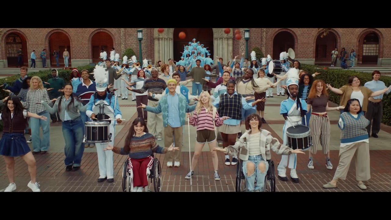 A diverse group of people, including a marching band and individuals in wheelchairs, smile and pose energetically in front of a building with arched windows.