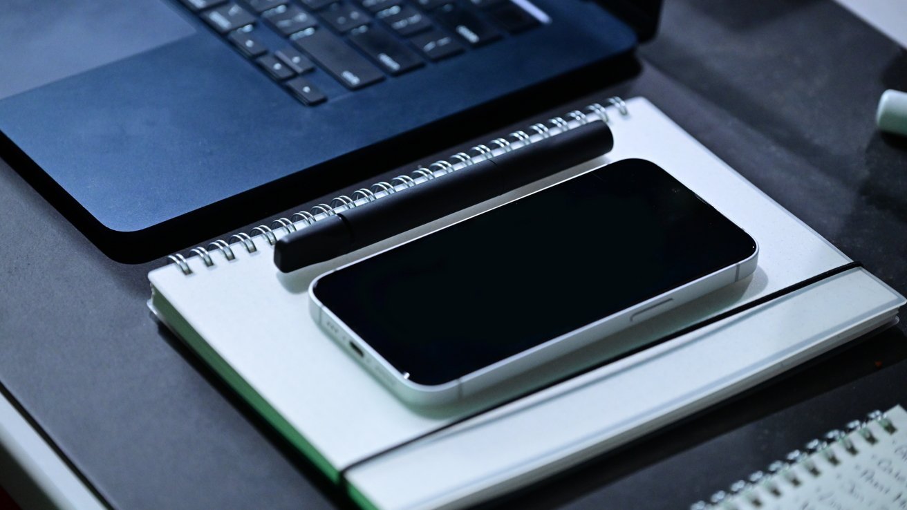 A smartphone, notebook, and pen are placed on a desk next to a laptop.