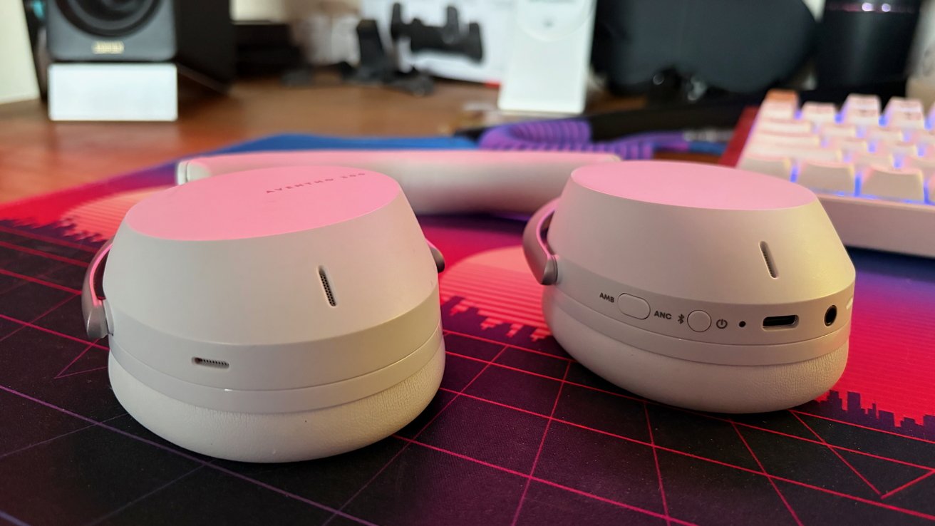 Two beige wireless headphones resting on a colorful patterned mat, with a keyboard and speaker visible in the blurred background.