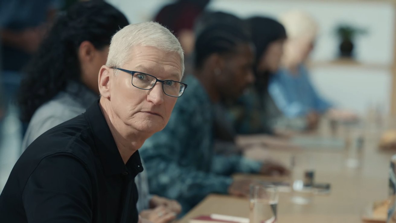 A man with glasses and gray hair in a black shirt sits at a conference table, with several people blurred in the background.