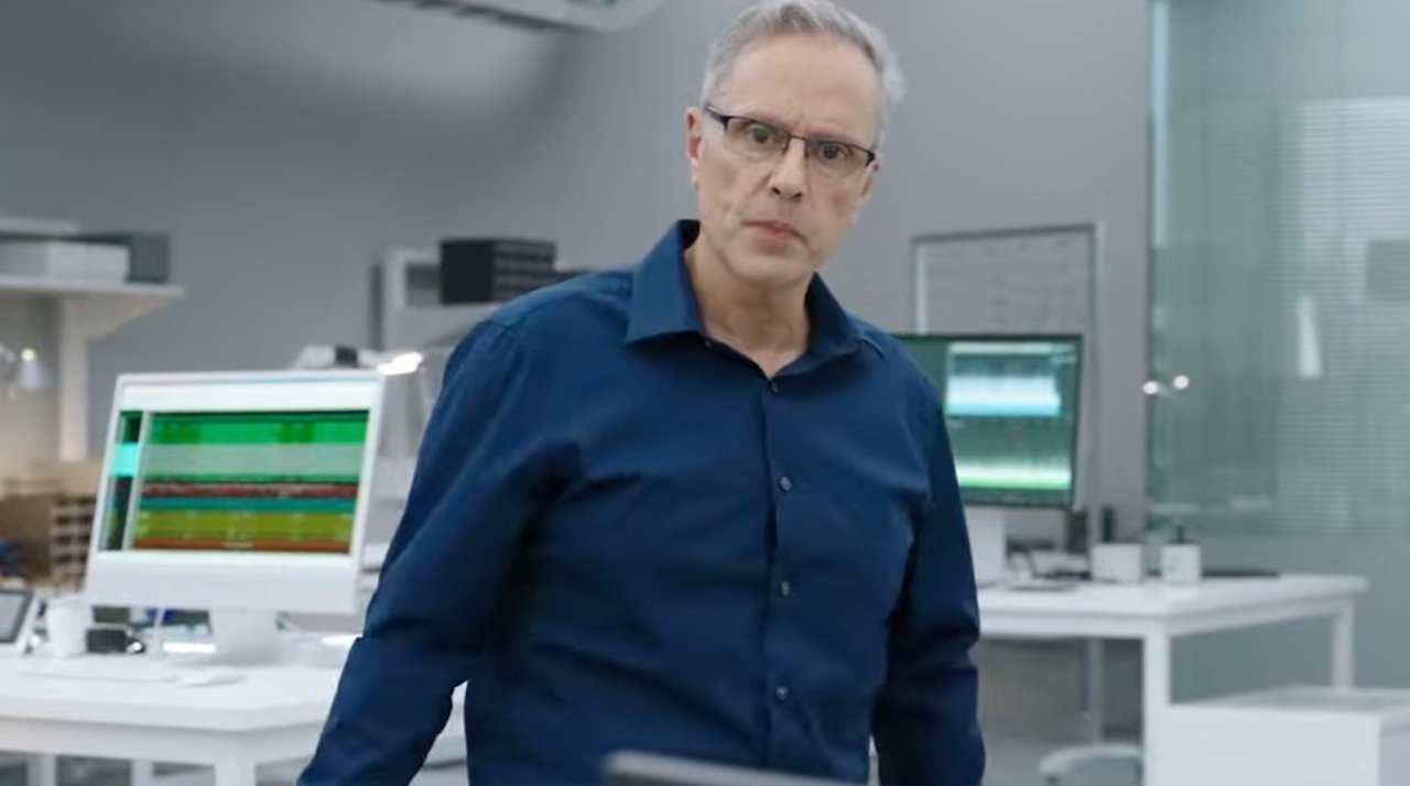Man in a blue shirt standing in a modern office with computers displaying data and white desks in the background.