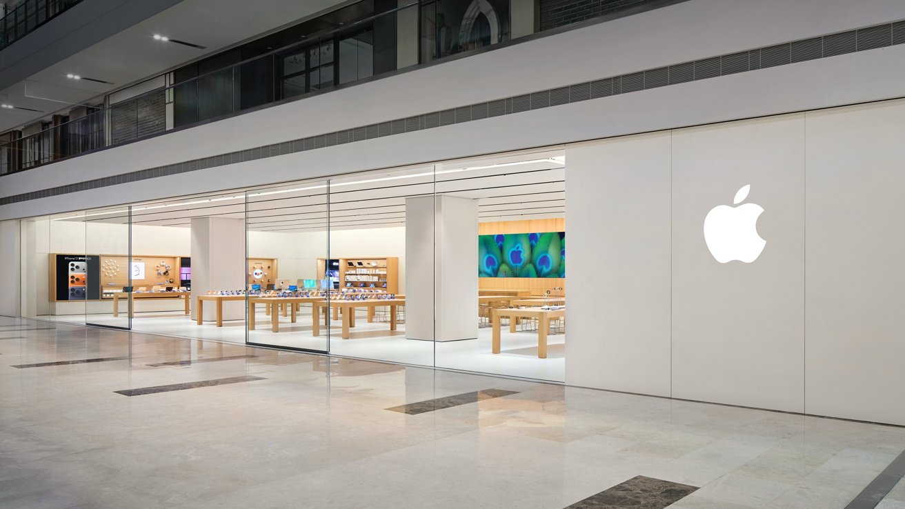 Apple store with glass walls, displaying tables of electronic products, wooden shelves, and a colorful peacock feather-themed art piece.