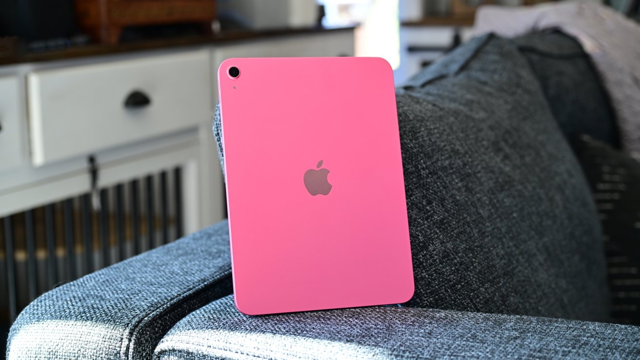 Pink tablet with a silver apple logo rests on a gray fabric couch in a softly lit room.