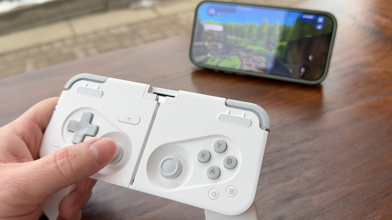 Hand holding a white gaming controller in front of a smartphone displaying a video game on a wooden table.