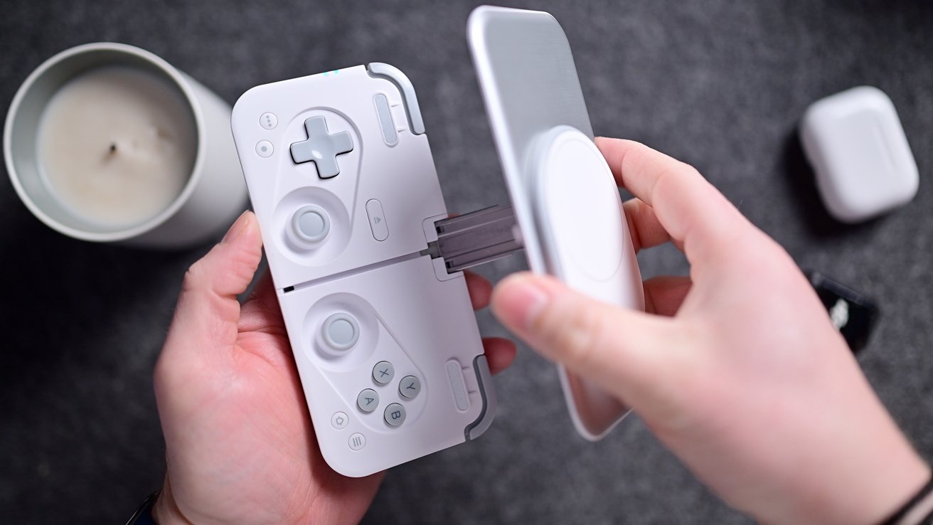 Person holding a white gaming controller case in both hands, with a smartphone being placed inside. A candle and earbud case appear in the background.