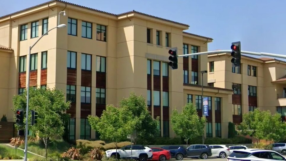 A modern, multi-story beige building with large windows, surrounded by trees and parked cars, beneath a clear blue sky and traffic lights.