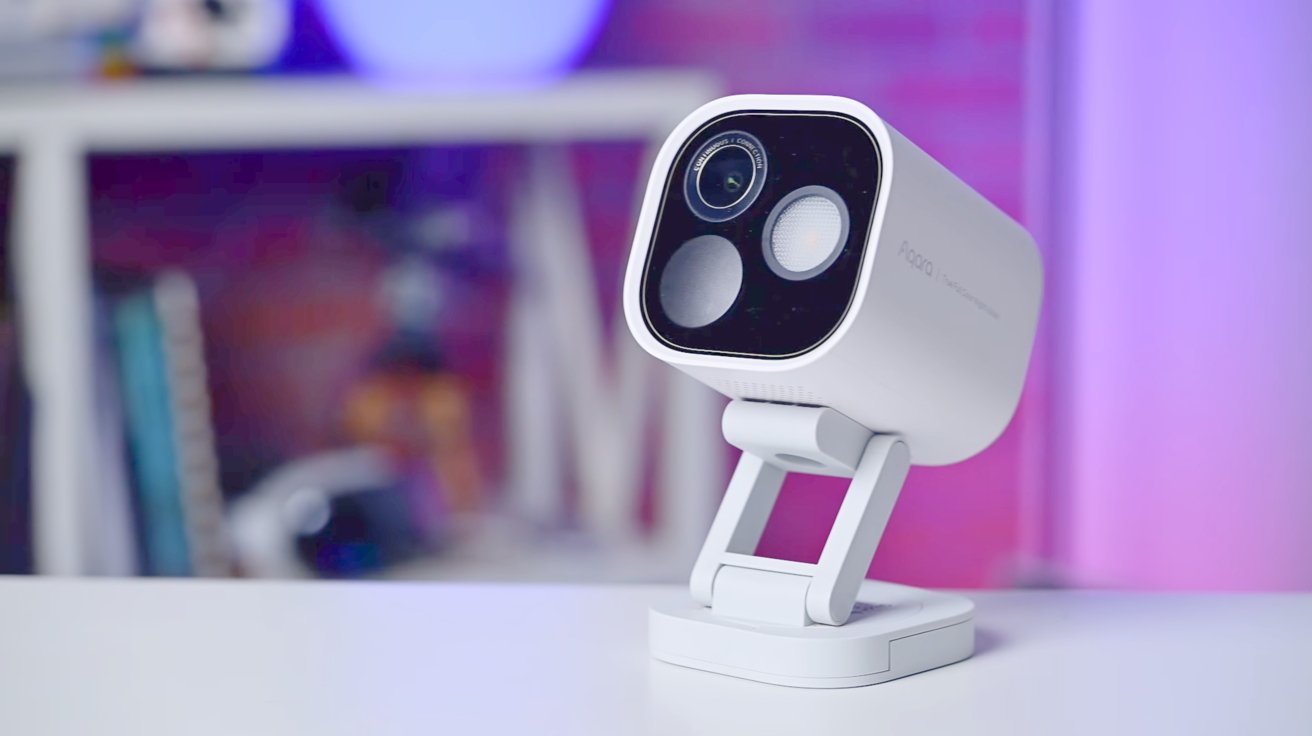 A modern white security camera with three lenses, mounted on an adjustable stand, against a blurred background with blue and pink lighting.