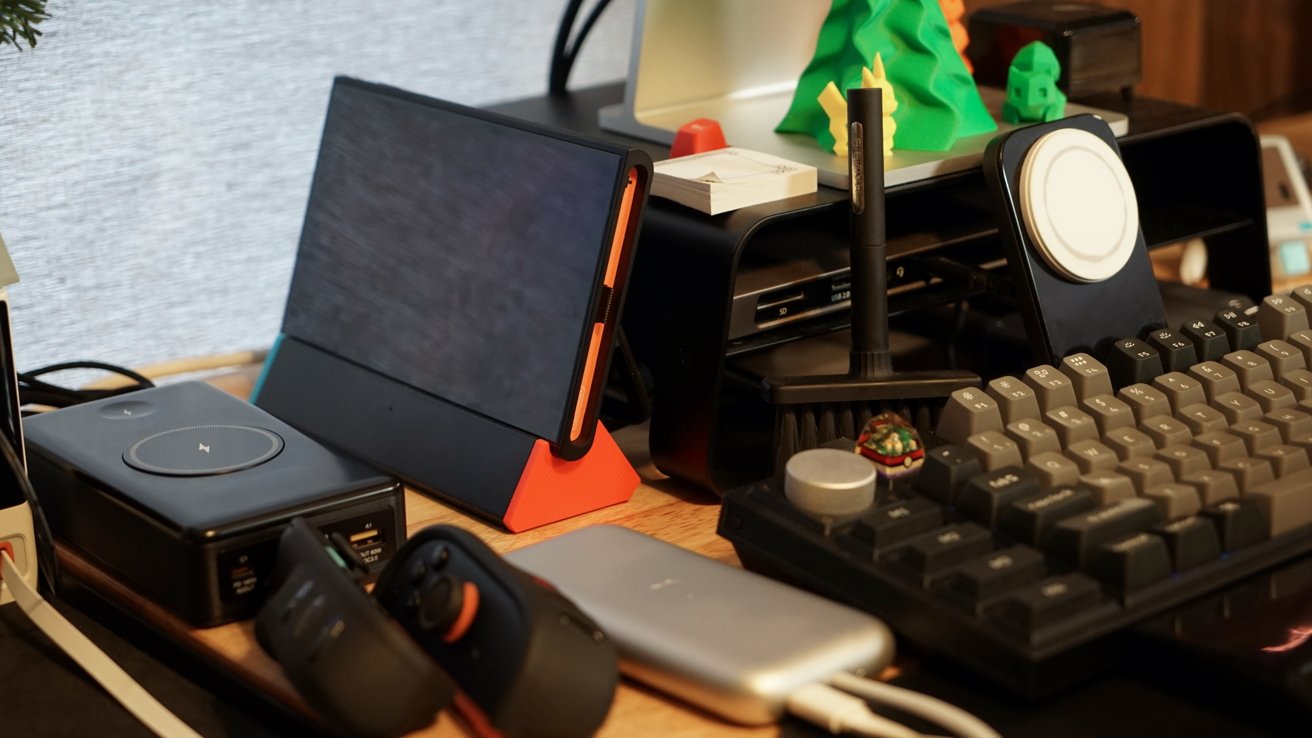 A desk with a tablet on a stand, a wireless charger, a keyboard, a gaming controller, and a small decorative tree and figurine.