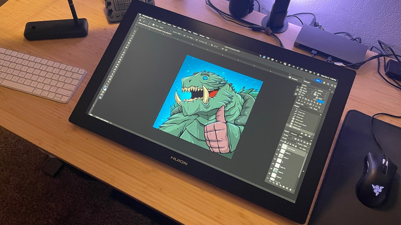 Graphics tablet displaying digital illustration of a cartoon monster giving a thumbs-up, with a computer keyboard and mouse on a wooden desk.