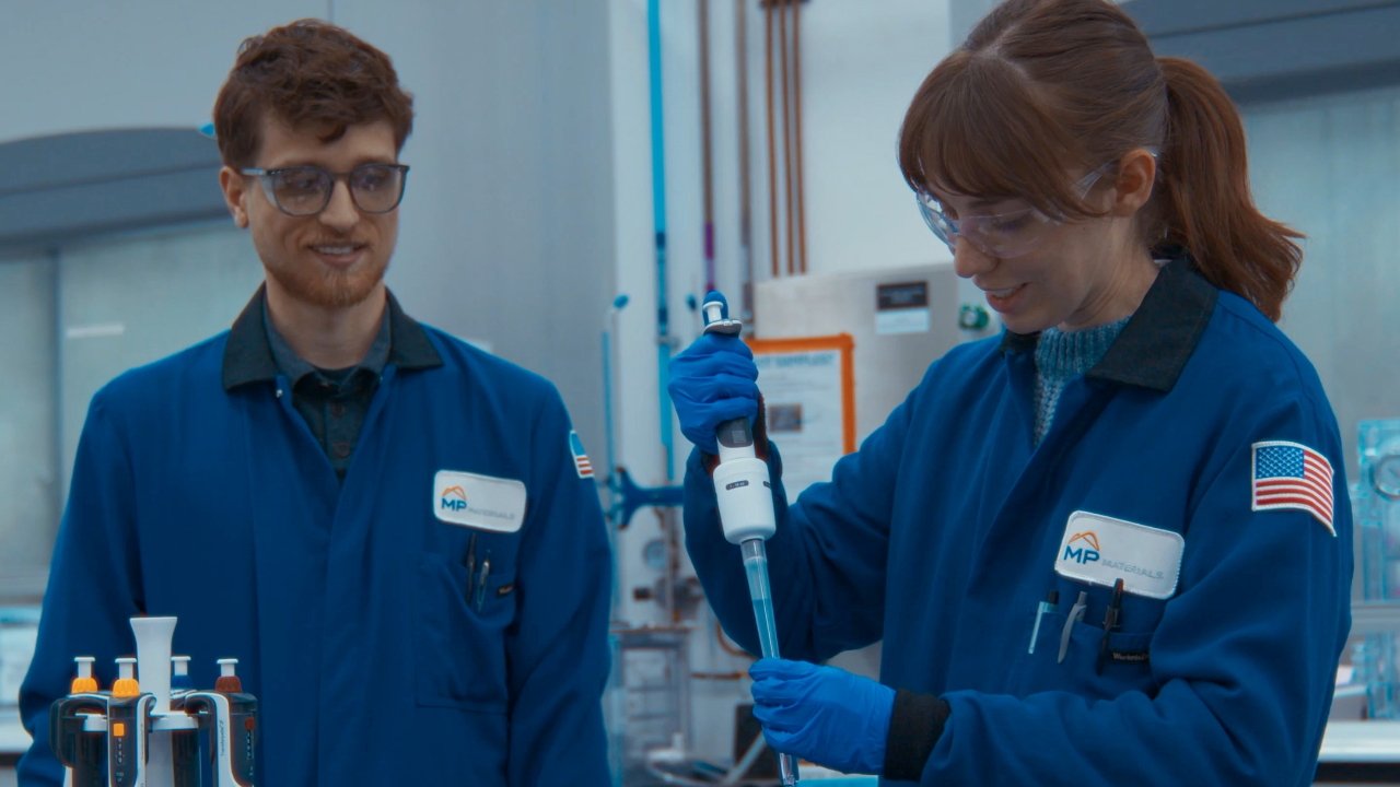 Two scientists in blue lab coats working with lab equipment, one using a pipette, both wearing safety glasses and gloves, smiling in a laboratory setting.