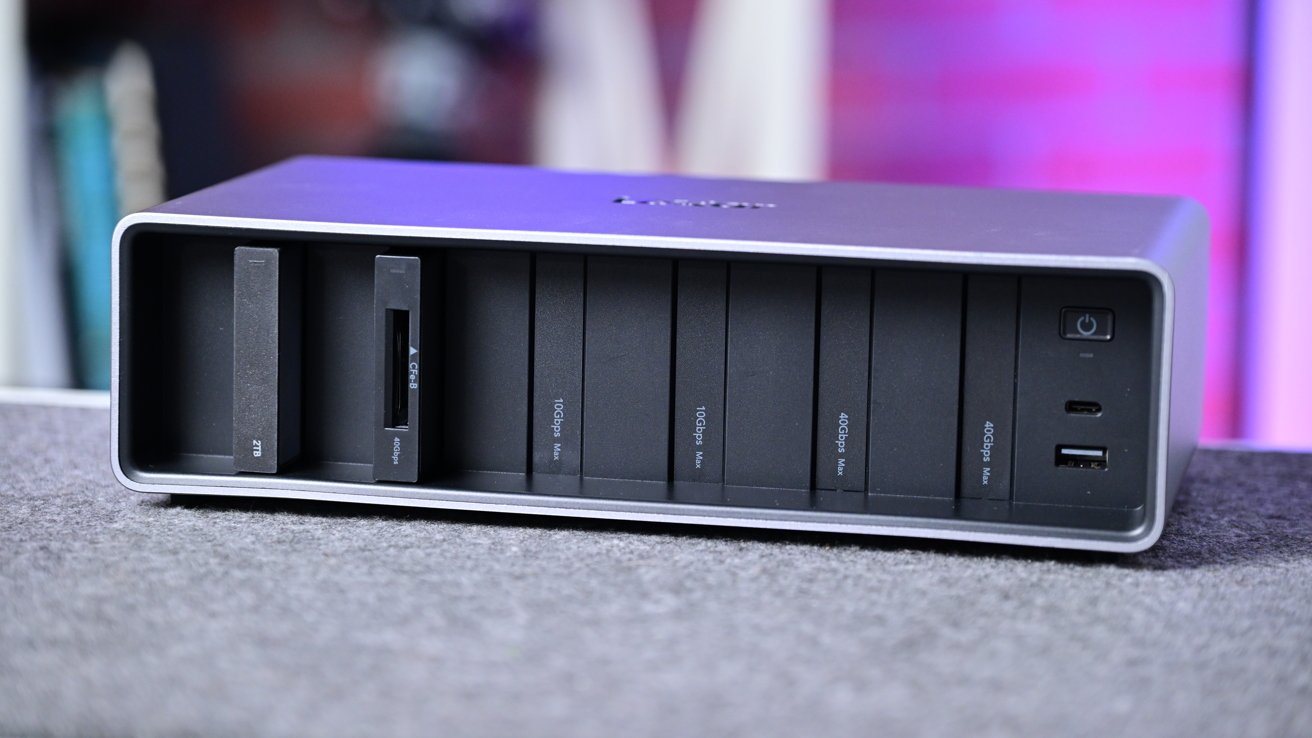 Black multi-slot device on a shelf with card reader, ports, and power button, against a blurred colorful background.