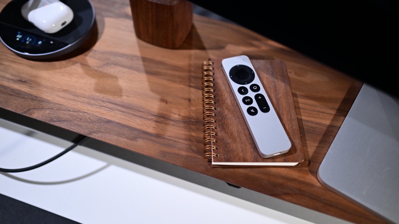Remote control on a spiral notebook atop a wooden desk, with a black circular device and white item nearby.