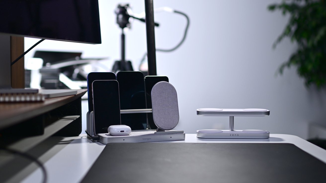 Charging station on desk with several smartphones, an earbud case, and wireless charging pads; blurred background with a monitor and plant.