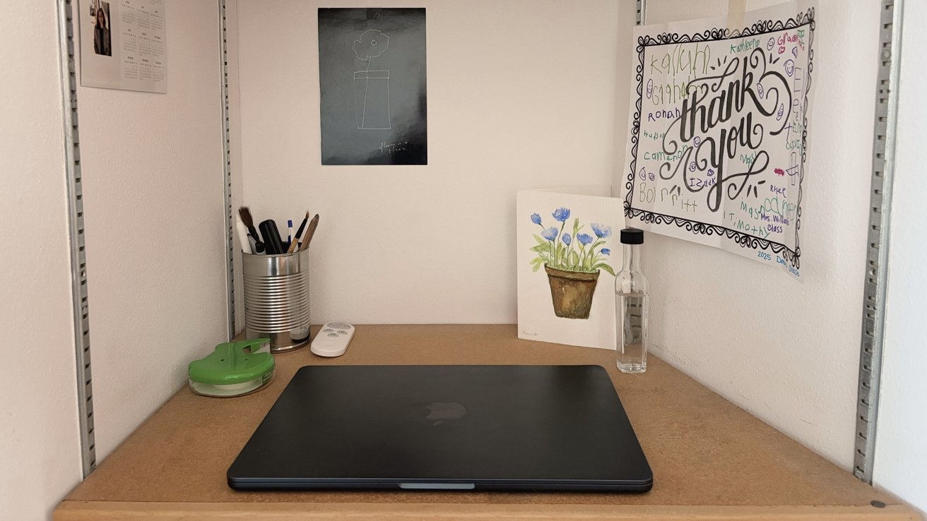 Desk with closed laptop, green tape dispenser, tin can holding pens and brushes, thank you card, watercolor painting, and small bottle. Wall displays a calendar and abstract drawing.