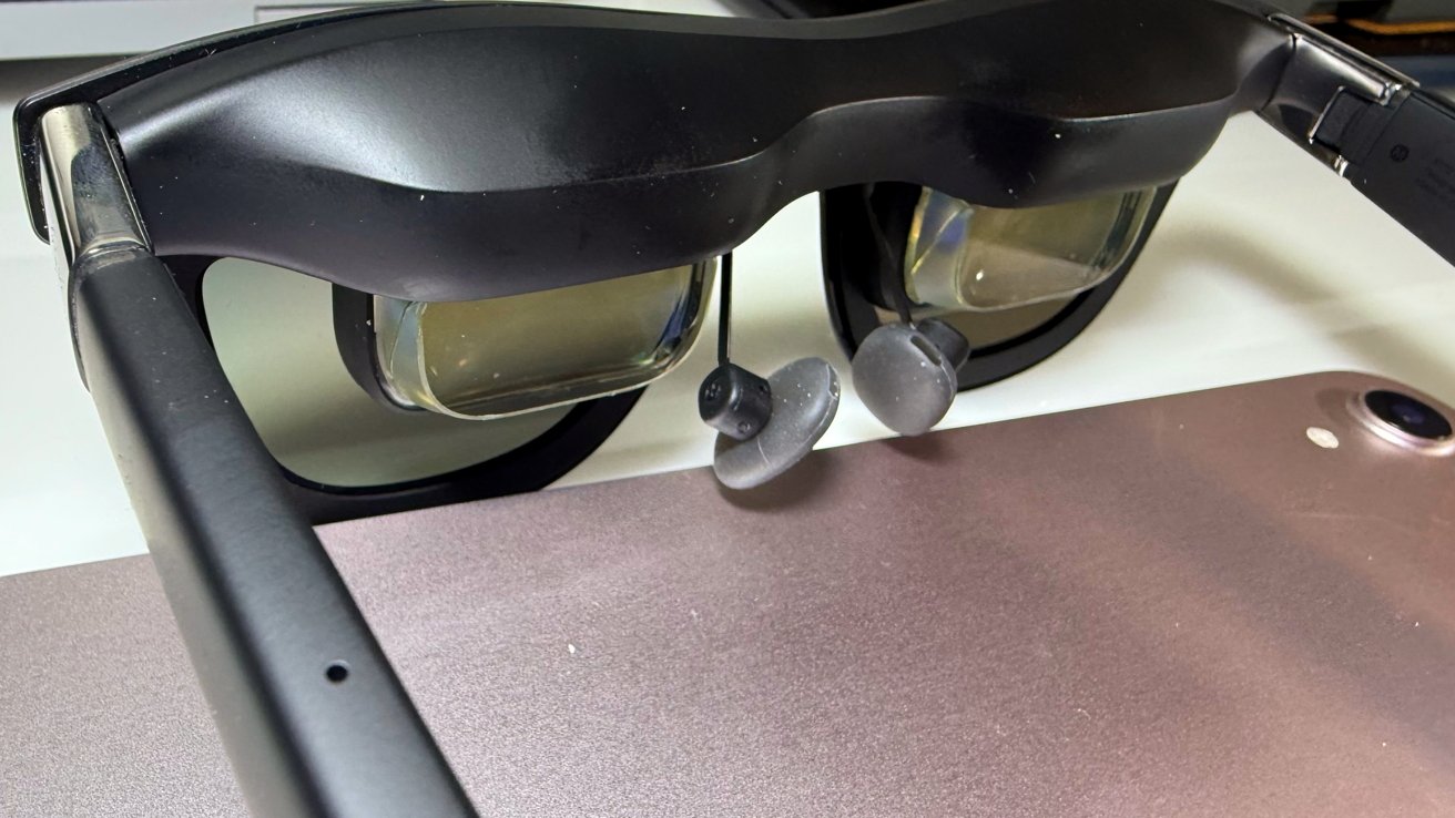 Black augmented reality glasses on surface with headphones on visible, next to metal tablet or device in background.