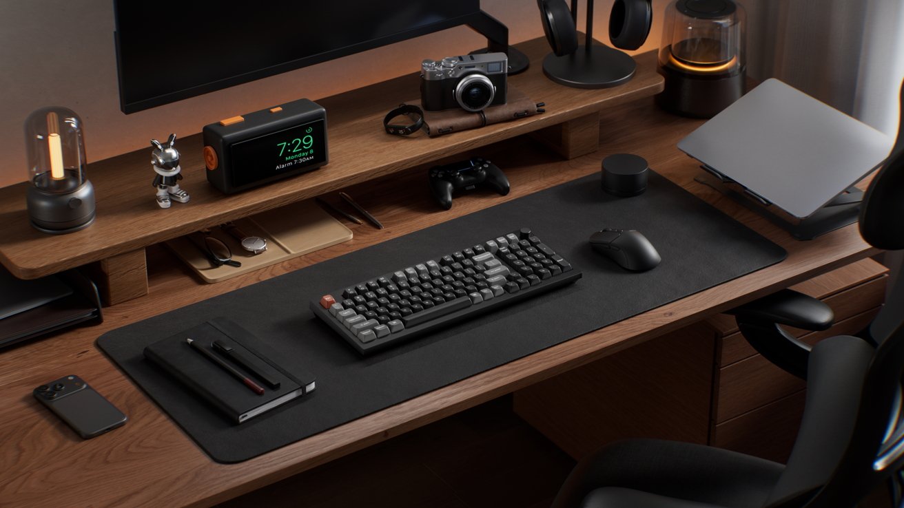 A wooden desk setup with a monitor, keyboard, mouse, black notebook, pens, clock, camera, headphones, lamp, and a phone, creating a neat, organized workspace.
