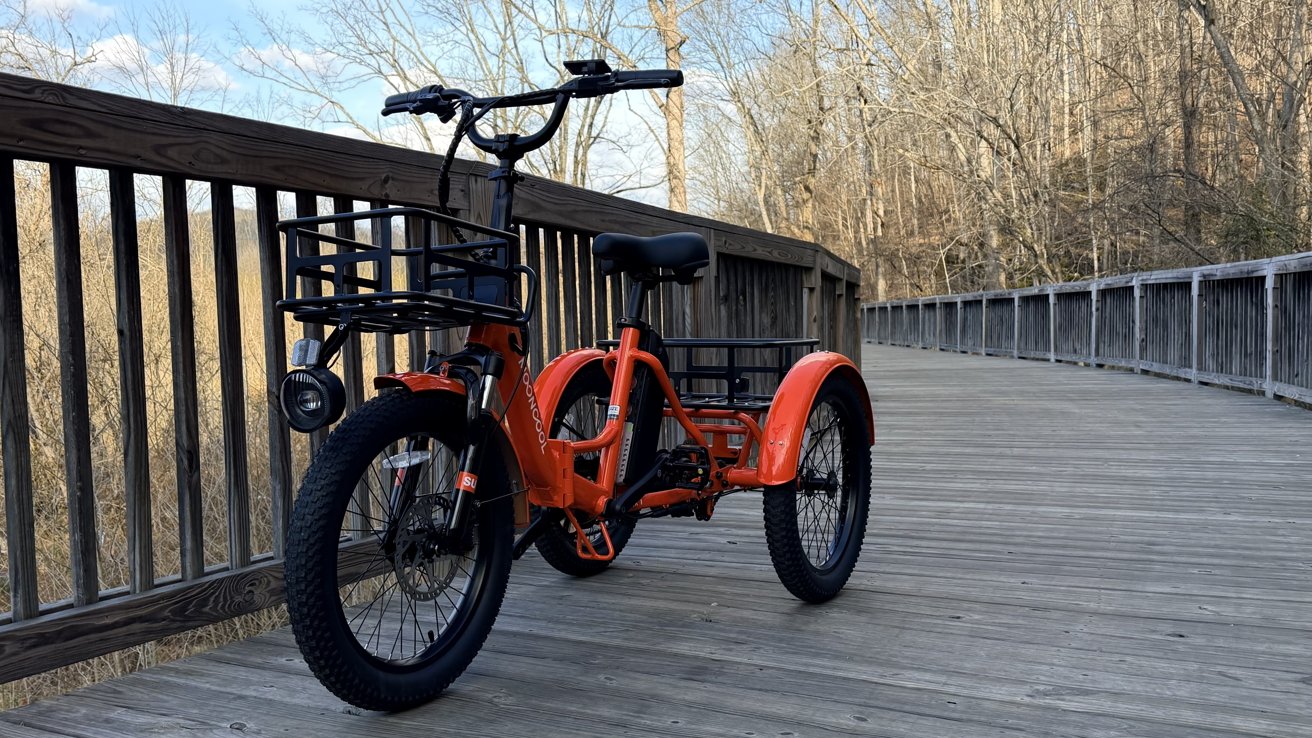 An orange tricycle with large wheels and a front basket stands on a wooden path, surrounded by leafless trees and wooden railings.