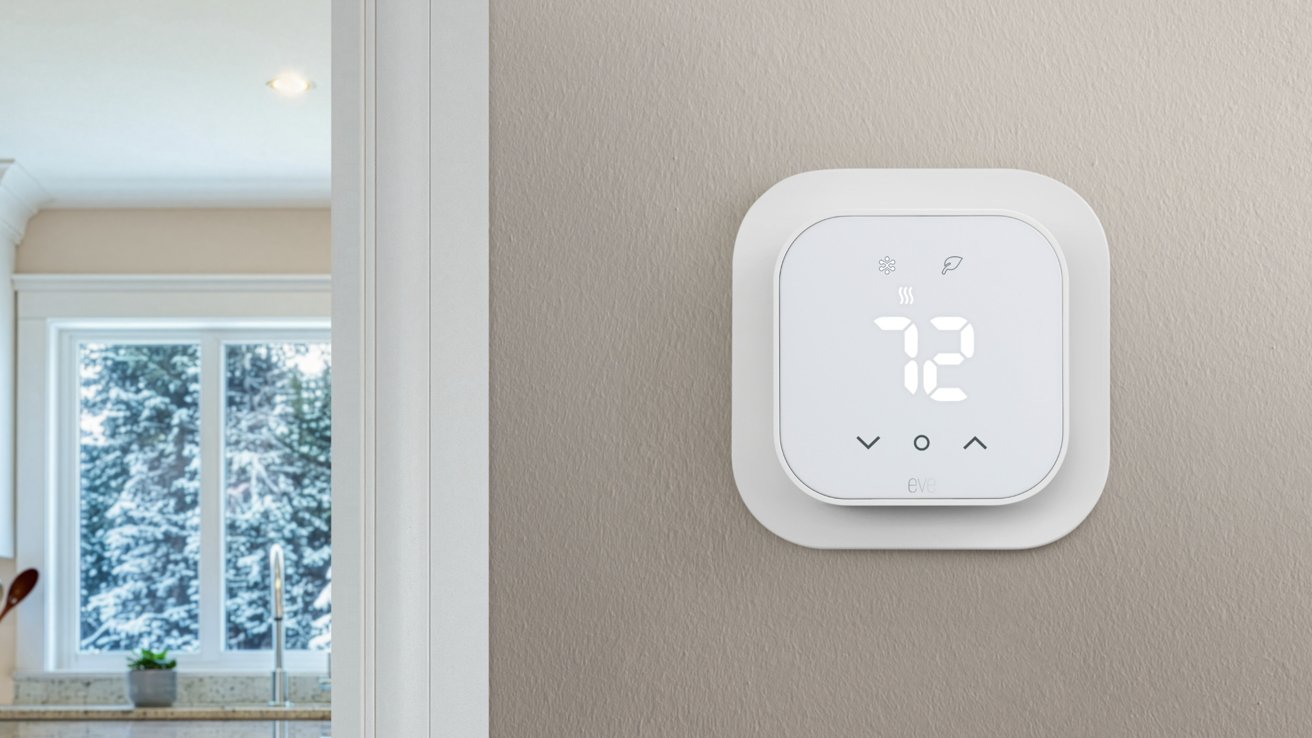 White smart thermostat on a beige wall displaying 72 degrees, with a kitchen window and snowy trees visible in the background.