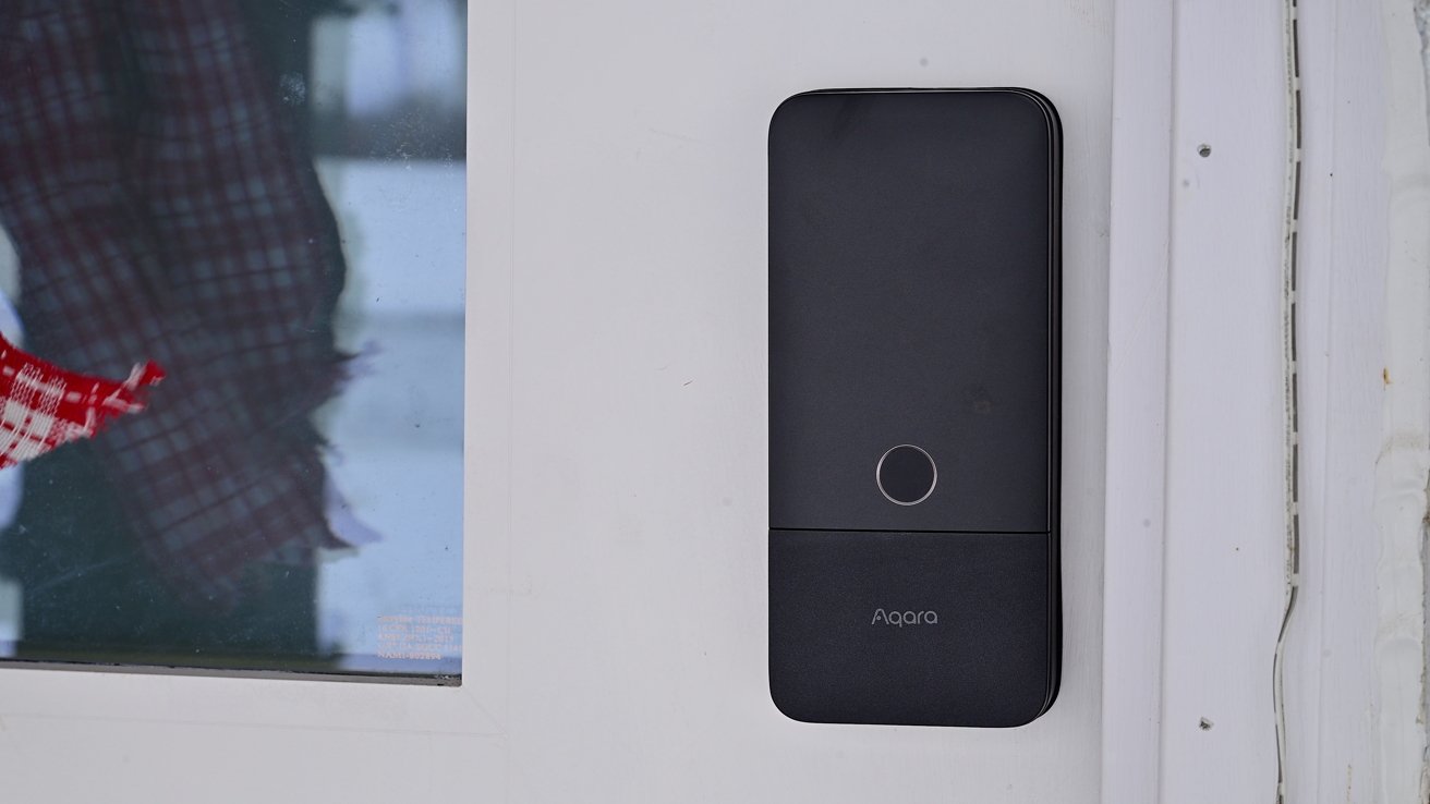 Black rectangular smart doorbell with a small circular button in the center, mounted on a white door frame next to a window showing a blurred person in plaid clothing