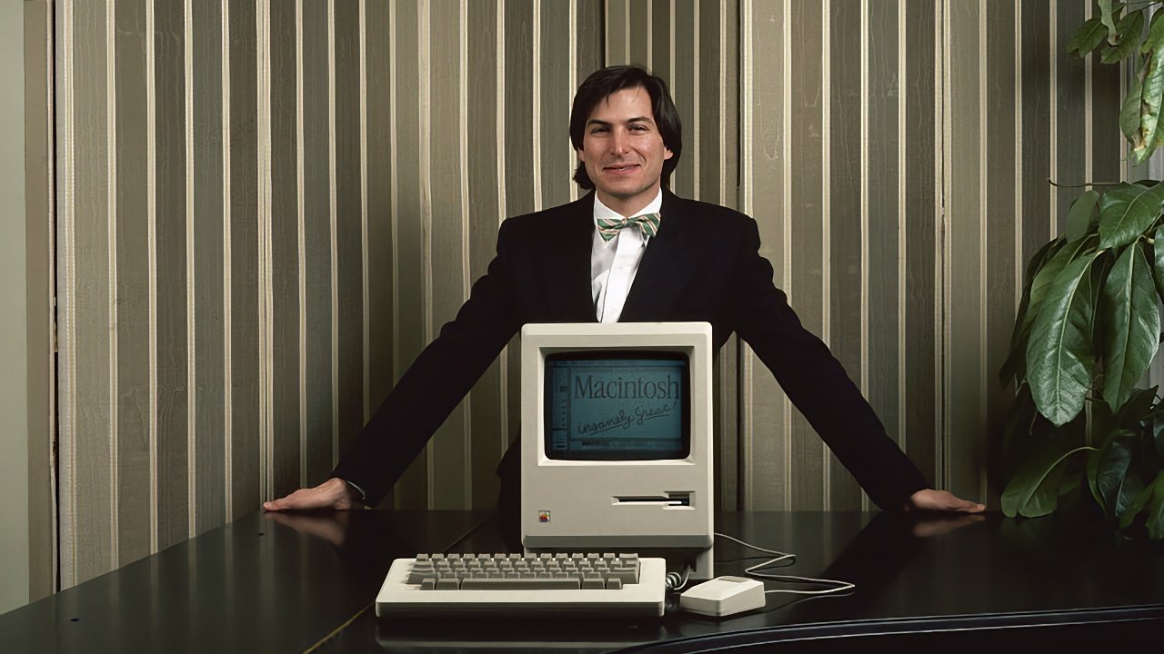 Man in a suit with a bow tie stands behind a vintage Macintosh computer on a dark table, striped wallpaper background.