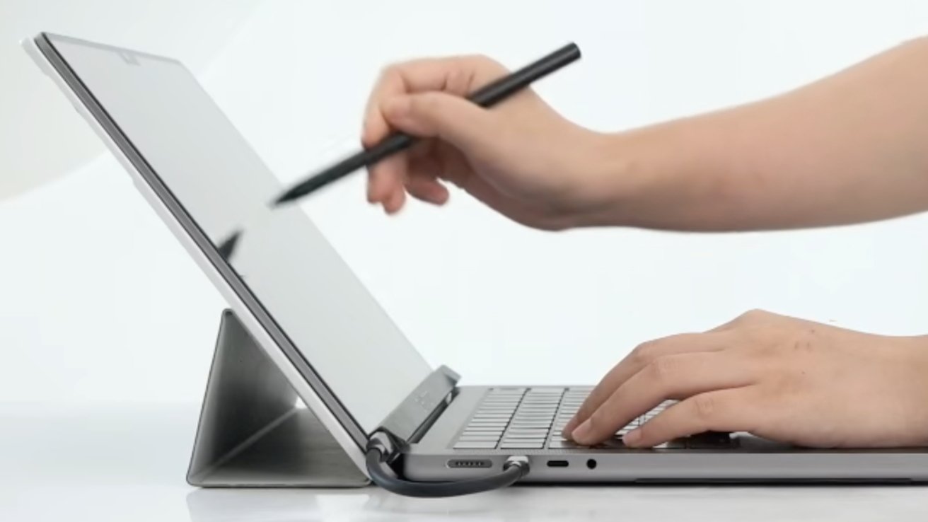 Hands using a stylus on a tablet stand and typing on a keyboard, with a cable connected to the device.