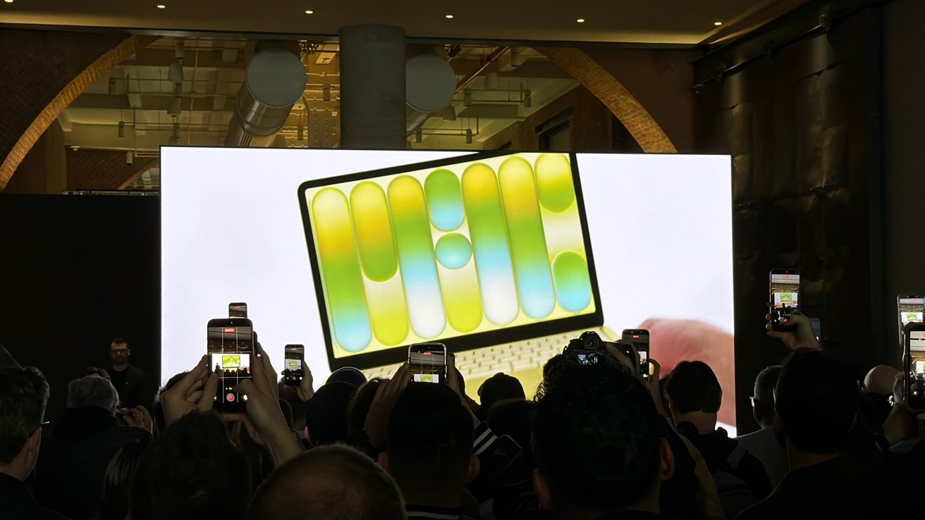 Crowd at a tech event photographing a large screen that displays a colorful tablet-like device with green, yellow, and blue abstract shapes on its screen.