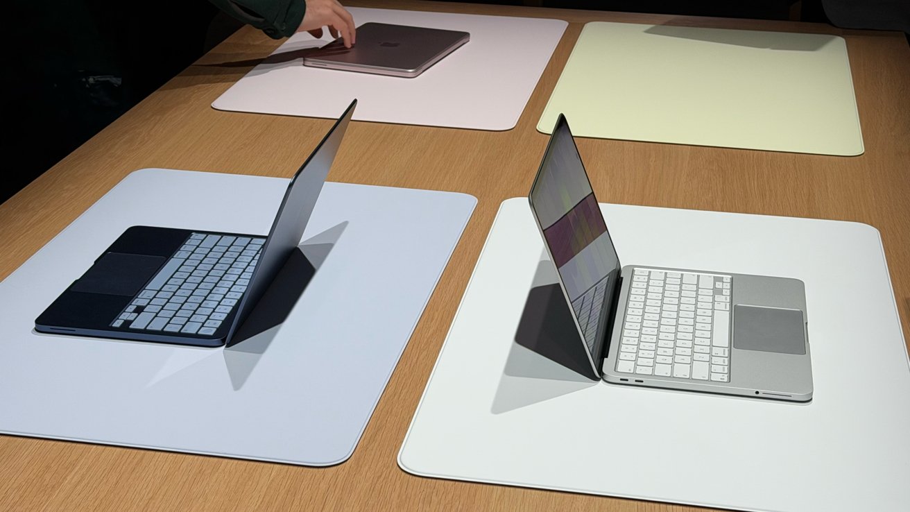 Two open laptops on large white mats atop a wooden table, screens angled upward, with a person's hand reaching toward a closed laptop in the background