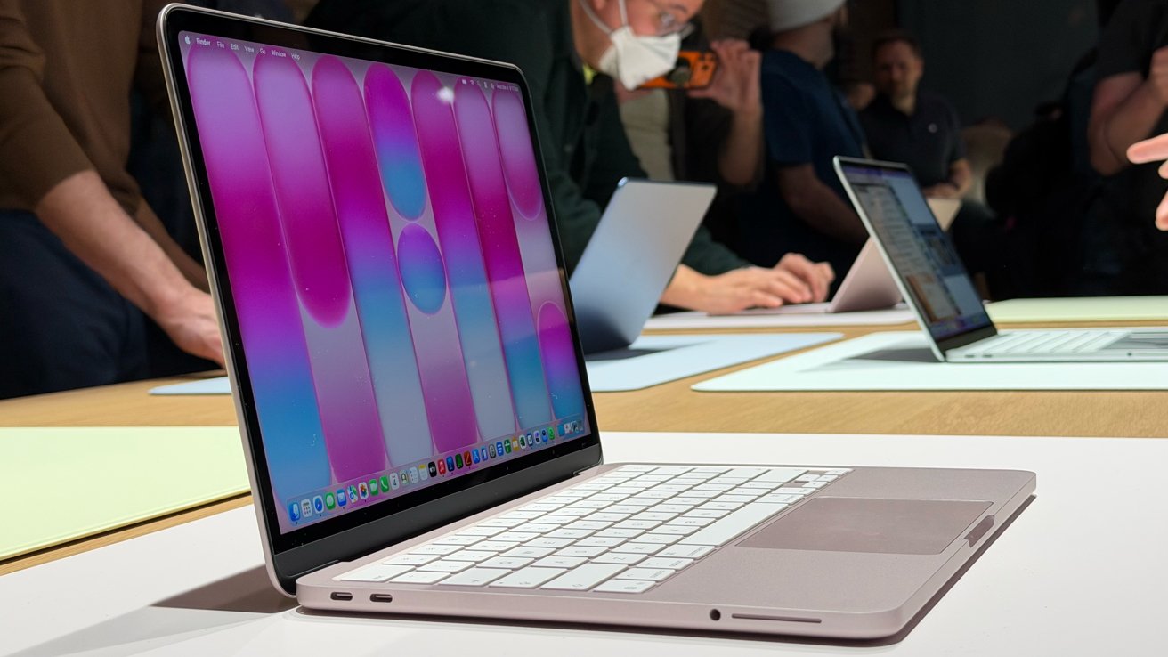Slim silver laptop on a table, screen showing colorful pink and blue abstract wallpaper, surrounded by other open laptops and people standing and working in a busy tech event setting