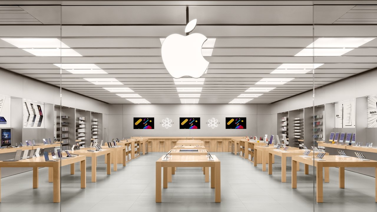 Bright, modern Apple retail store interior with large glowing Apple logo, wooden tables displaying laptops, tablets, and phones, wall shelves of accessories, and colorful product screens at the back