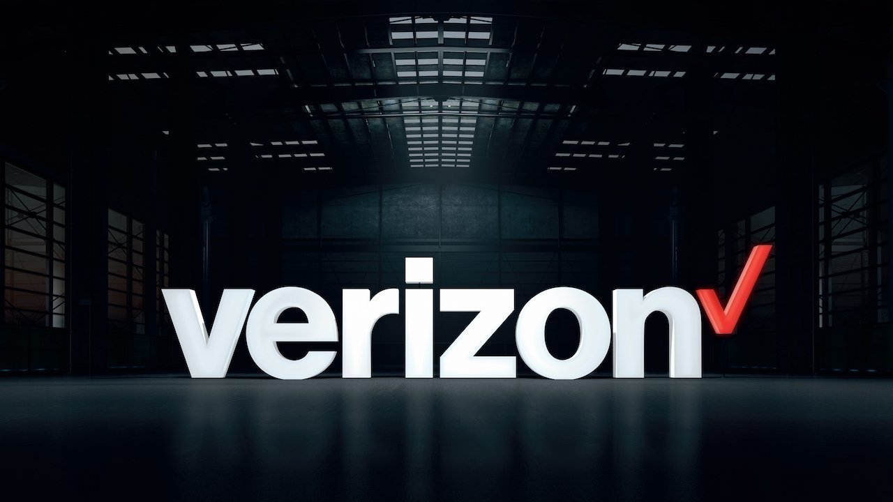 Large 3D white Verizon logo with a red checkmark at the end, centered on a dark, industrial warehouse background with overhead lights reflecting on the floor