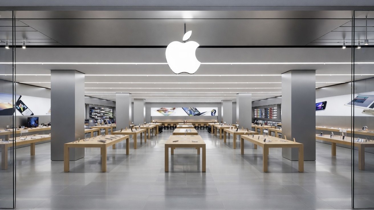 Spacious modern Apple store interior with large glowing Apple logo, sleek gray walls, bright ceiling lights, and rows of wooden tables displaying neatly arranged laptops, tablets, and phones