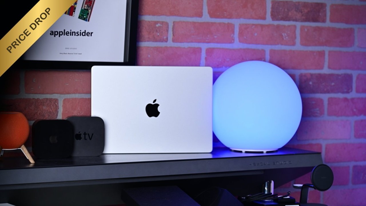 Silver MacBook Pro 14-inch with Apple logo on a desk, flanked by Apple TV boxes, orange smart speaker, glowing blue spherical lamp, and framed AppleInsider poster against a brick wall