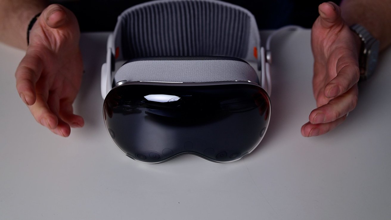 Hands presenting a sleek, dark-fronted virtual reality or mixed reality headset resting on a table, with a padded, ribbed head strap visible behind it