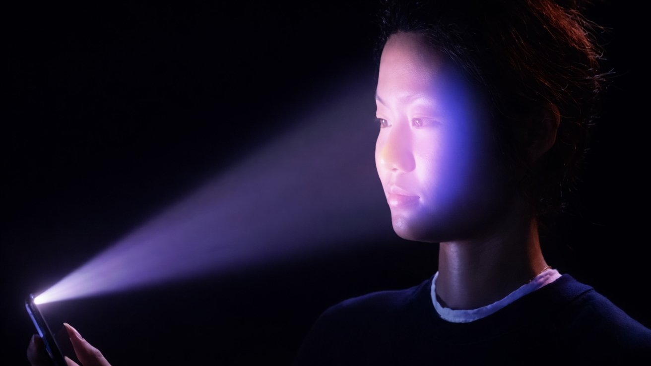 Person in a dark setting holding a smartphone, face illuminated by a bright purple-blue light beam from the screen, suggesting facial recognition or biometric scanning technology