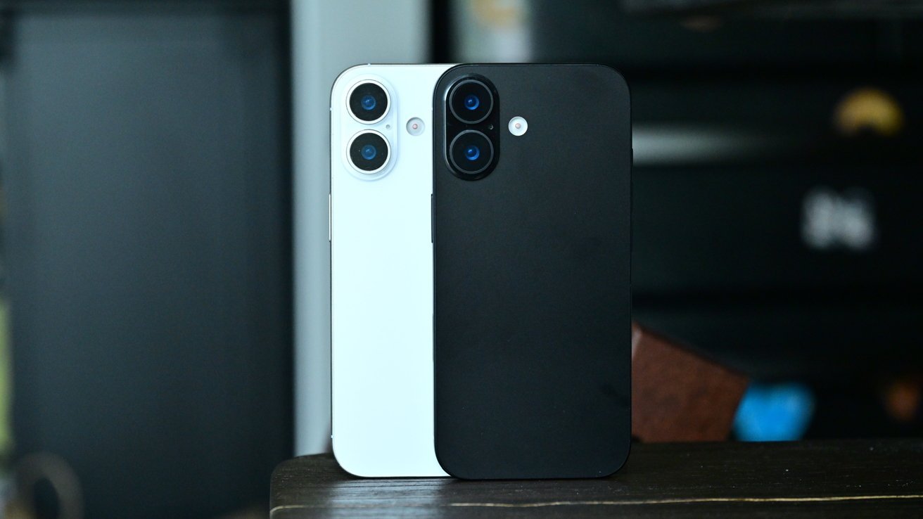 Two modern smartphones standing upright on a table, one white and one black, both showing their backs with dual rear cameras and flashes, against a softly blurred indoor background