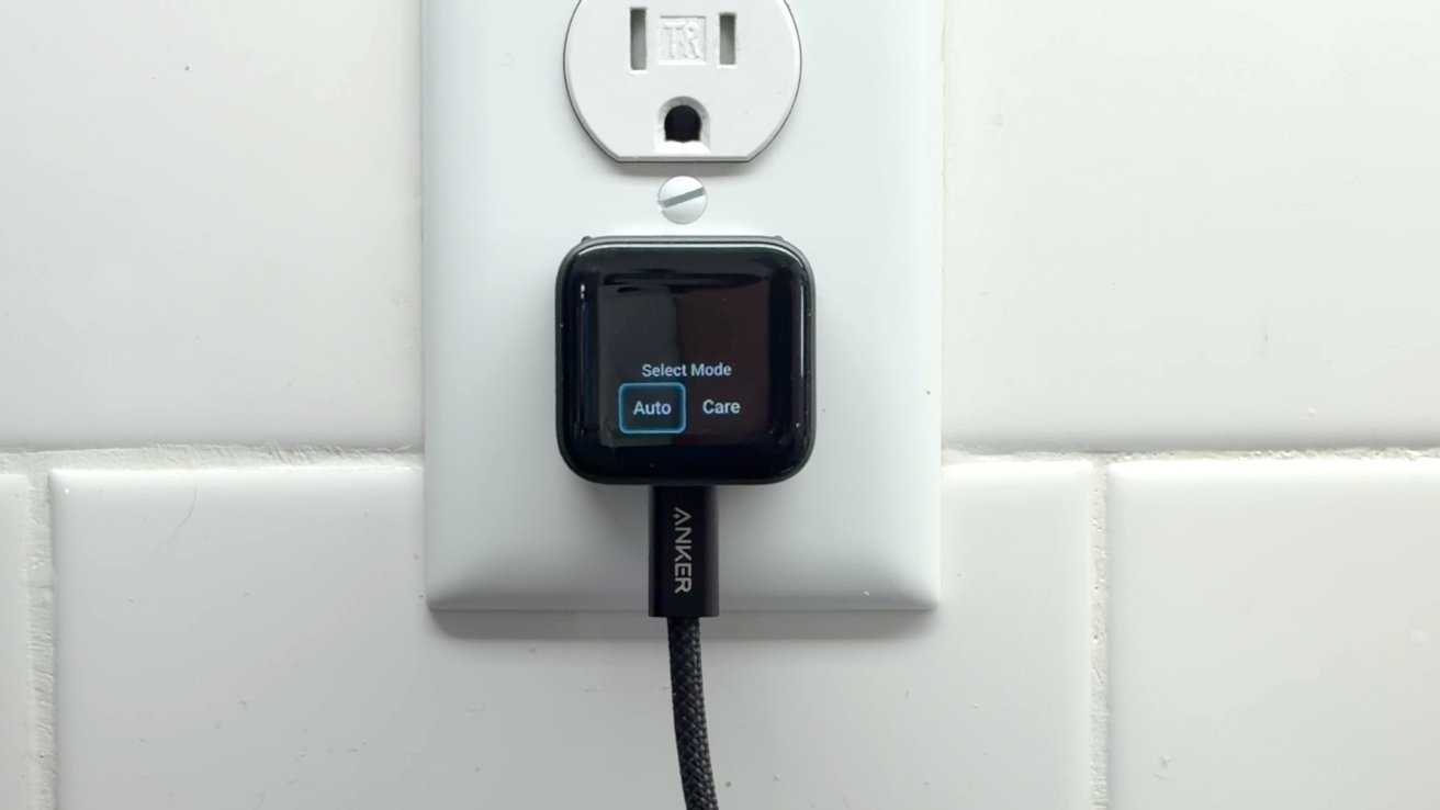 Small black Anker USB charger plugged into a white wall outlet above white tile, with a short screen displaying selectable modes labeled Auto and Care