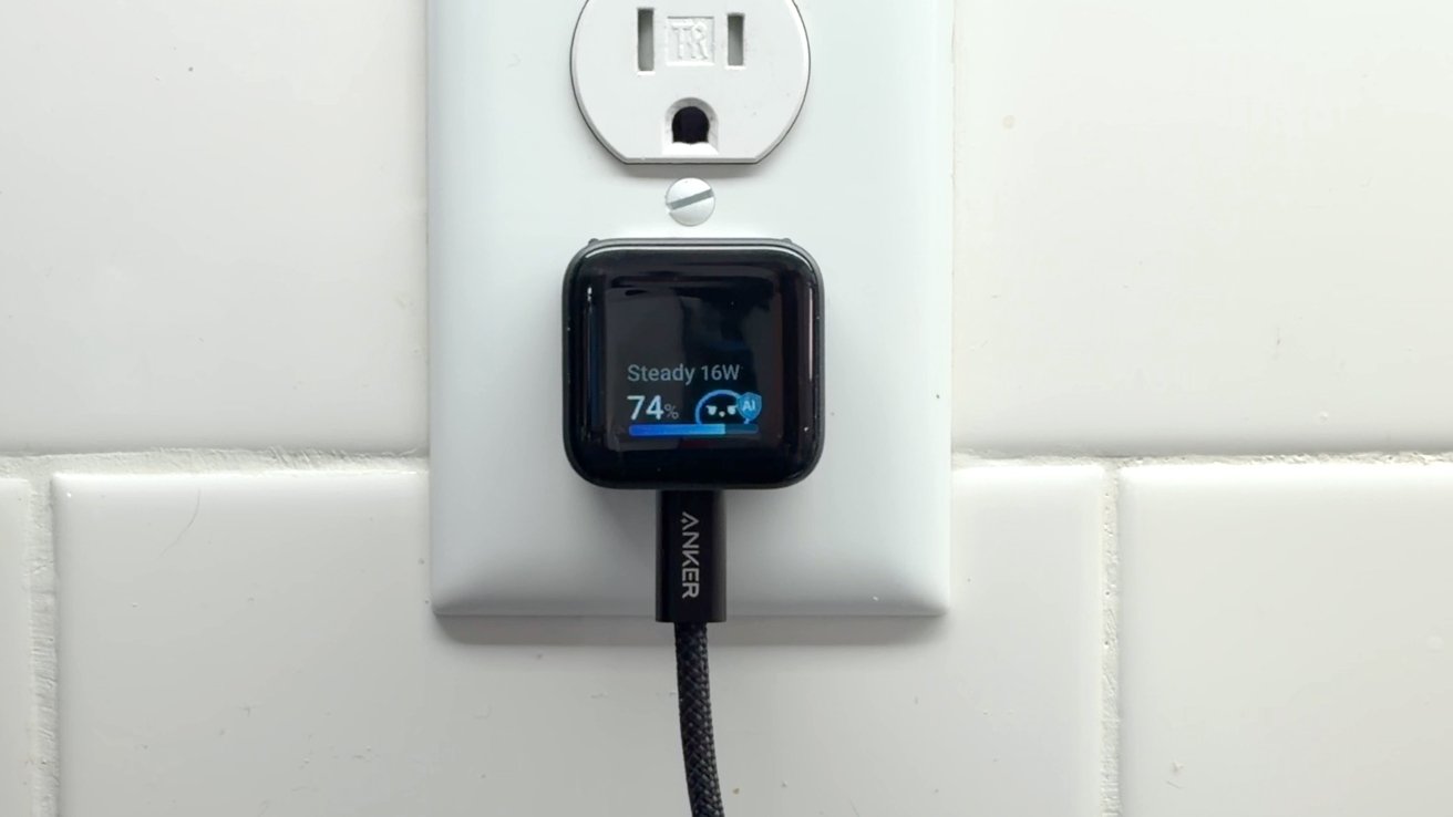 Small Anker wall charger plugged into a white electrical outlet, featuring a digital screen showing charging status at 74 percent, with a braided charging cable connected at the bottom