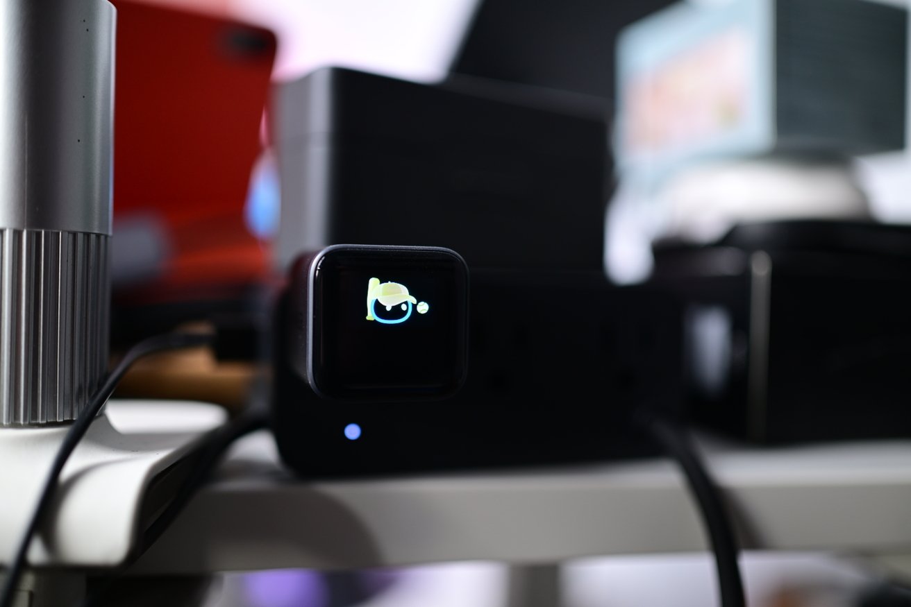 Small black electronic device with a glowing camera icon on its front, sitting on a cluttered white desk surrounded by blurred computer equipment and cables in the background