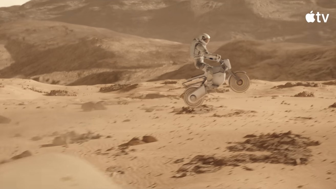 Astronaut in a white spacesuit rides a futuristic bike over dusty, cratered Martian-like desert terrain, catching air amid rolling hills under a hazy brown sky, Apple TV logo in corner