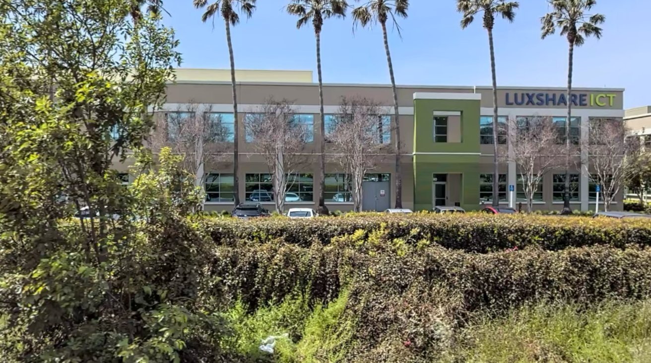 Two-story Luxshare ICT office building behind tall palm trees and parked cars, viewed across a dense green hedge and shrubs under a bright blue sky.