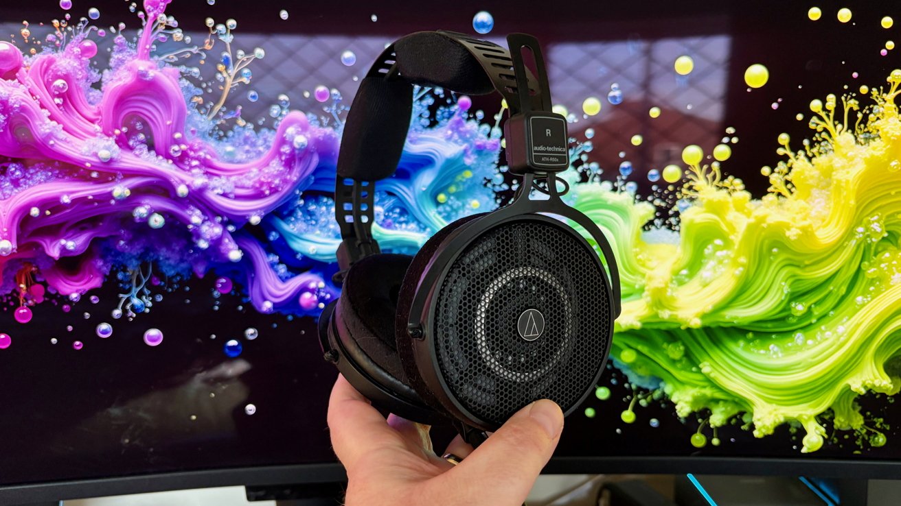 Hand holding large black Audio-Technica over-ear headphones in front of a computer monitor displaying vibrant swirling splashes of pink, purple, blue, green, and yellow paint-like colors