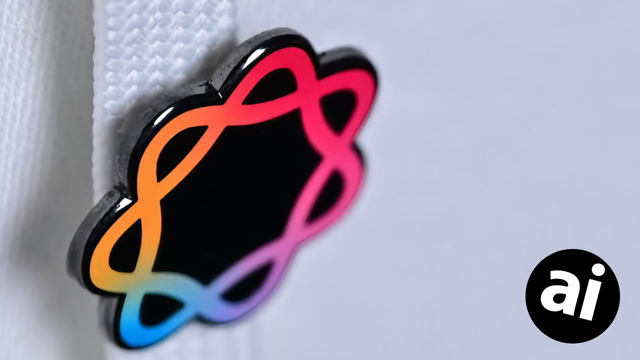 Colorful looping flower-shaped pin with black center attached to white textured fabric, with a small black circle logo containing lowercase letters ai on a pale background