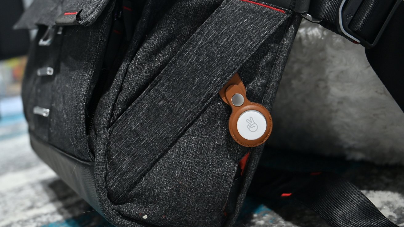 Dark gray backpack with a small round tracking tag in a brown holder clipped to a side pocket, resting on a patterned surface indoors